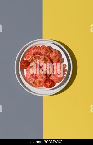 Blick von oben auf eine leuchtend rote raf-Tomate, die in zwei Hälften geschnitten, mit Salz und Gewürzen garniert und auf einem weißen Teller platziert wird. Der gelbe und homosexuelle Hintergrund hebt ihn hervor Stockfoto