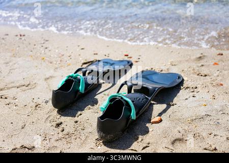 Am Sandstrand in der Nähe des Wasserrandes liegen schwarze Schwimmflossen mit blauen Riemen und sanften Wellen am Ufer, die auf einen Tag hindeuten Stockfoto