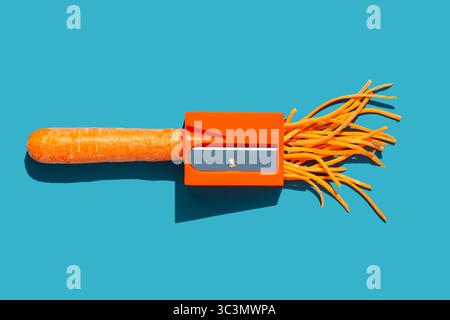 Draufsicht einer orangefarbenen Karotte in einem orangefarbenen Bleistiftspitzer auf einem leuchtend blauen Hintergrund. Die frisch rasierten Karottenlocken sorgen für einen verspielten Touch Stockfoto