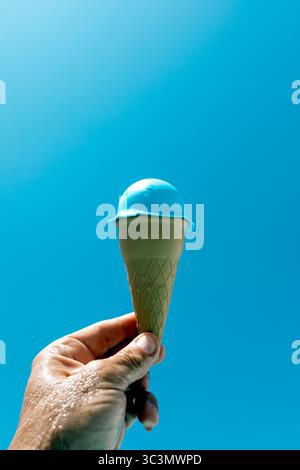 Eine abgeschnittene, nicht erkennbare Hand für Erwachsene, die eine blaue Spielzeugeiskegel vor einem klaren blauen Himmel hält, sorgt für einen verspielten Sommer-Vibe mit leuchtenden Farben und einem Stockfoto