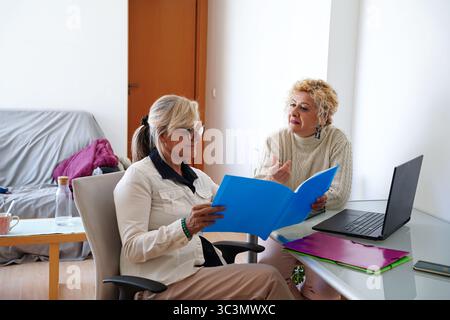 Zwei hochrangige Freundinnen werden in einem gemütlichen Zuhause gesehen, während sie sich bei der Lektüre von Dokumenten an einer durchdachten Diskussion beteiligen. Eine Frau hört aufmerksam zu Stockfoto