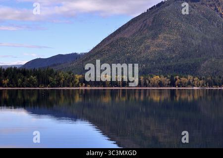 Goldene Herbstwälder säumen die stillen Gewässer des Lake McDonald und spiegeln schroffe Berghänge und nebelige Himmel in einem ruhigen Panorama der Wildnis von Montana wider. Stockfoto