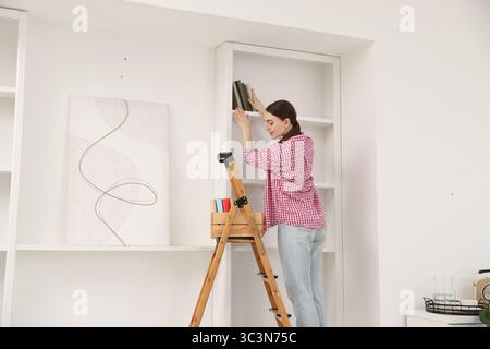 Frau, die zu Hause Bücher in Regale legt, indem sie Leiter benutzt Stockfoto