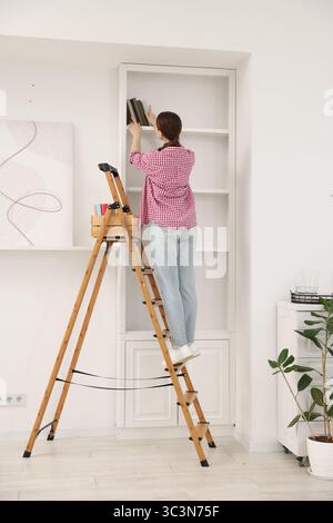 Frau, die zu Hause Bücher in Regale legt, indem sie Leiter benutzt Stockfoto