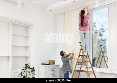 Ein Paar, das Vorhänge mit einer Leiter im Zimmer aufstellt Stockfoto