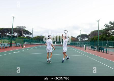 Verschiedene männliche Tennispartner in weißen Outfits feiern den Sieg auf dem grünen Platz mit Schlägern und Netz Stockfoto