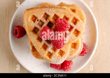 Detaillierte Nahaufnahme goldener herzförmiger Waffeln mit Puderzucker und lebendigen frischen Himbeeren auf einem weißen gesprenkelten Teller Stockfoto