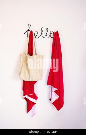 Zwei rote Strandtücher und eine Tasche hängen an einem Kleiderbügel in einem Hotel. Der Aufhänger ist in Form der Inschrift Hallo. Handtücher gegen eine weiße Wand. Stockfoto