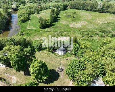 Ein Blick aus der Vogelperspektive auf eine kleine Kapelle, umgeben von grünen Bäumen, einem sich windenden Fluss und offenen Feldern unter hellem Himmel Stockfoto