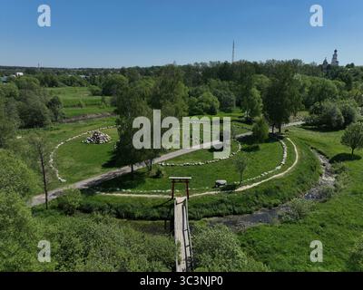 Ein Panoramablick auf einen malerischen Park mit runden Wanderwegen, üppigen grünen Bäumen, einer kleinen Brücke und einer weit entfernten Kirche Stockfoto