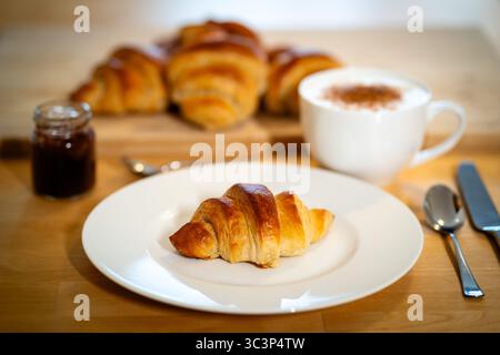 Frisch gebackenes goldbraunes Croissant auf einem weißen Teller, begleitet von einer Tasse Cappuccino und einem Glas Marmelade Stockfoto