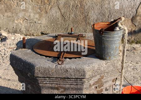 Ein verwitterter Steinbrunnen steht im Freien, gekrönt mit einem rostigen Metalldeckel und einem alten Blechkübel mit Seil, das traditionelles ländliches Wasser repräsentiert Stockfoto