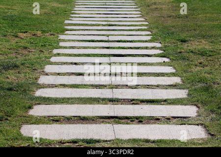 Rechteckige Betongesteine, die gleichmäßig in einer geraden Linie angeordnet sind, bilden einen Weg durch einen üppigen grünen Rasen und erzeugen einen visuellen Rhythmus in einer Landschaft Stockfoto