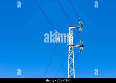 Ein harter weißer Metallpylon steht vor einem leuchtend blauen Himmel und unterstützt mehrere Stromleitungen mit Glasisolierungen und einem kleinen Vogel-perc Stockfoto