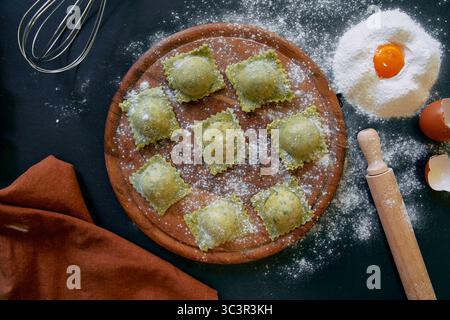 Hausgemachtes grünes Ravioli gefüllt mit frischen Zutaten auf einem Holzbrett, bereit zum Kochen zwischen Mehl und Eierbestandteilen Stockfoto