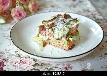 Köstliches Sandwich mit offenem Gesicht, zartem Fleisch, knusprigem Gemüse und herzhaftem Käse, serviert auf einer Tischdecke mit Blumenmuster Stockfoto