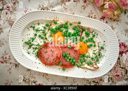 Ein köstlich gesunder Frühstücksteller mit Eiern, Schinken und frischen Grünen Erbsen, serviert auf einer Tischdecke mit Blumenmuster Stockfoto