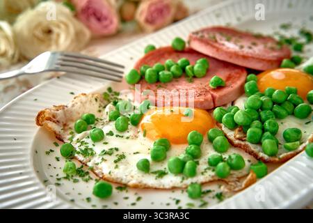 Köstlicher Frühstücksteller mit sonnigen Eiern, geschnittenem Schinken und frischen grünen Erbsen, die auf einem Deko-Teller liegen und von wunderschöner Blüte umgeben sind Stockfoto