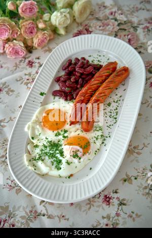 Ein köstlicher Teller mit gebratenen Eiern, Würstchen und roten Kidneybohnen, garniert mit Kräutern auf einer Blumentischdecke Stockfoto