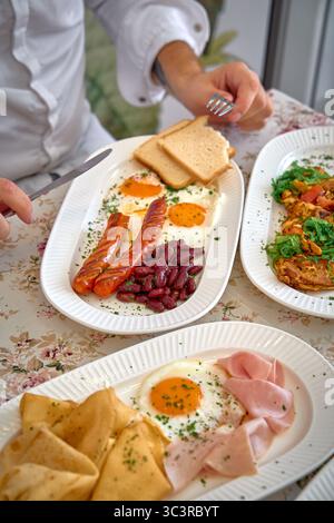 Ein köstliches Frühstücksangebot mit Eiern, Würstchen, Kidneybohnen, Schinken und Crepes auf einem floralen Tischtuch für ein köstliches Frühstück Stockfoto