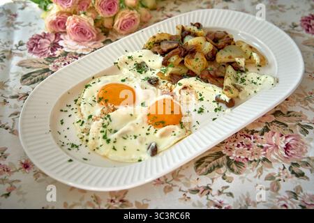 Köstlicher Frühstücksteller mit zwei Eiern auf der sonnigen Seite, garniert mit cremiger Sauce, serviert mit gewürzten Kartoffeln und frischen Kräutern im floralen Deko Stockfoto