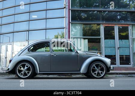 Bukarest, Rumänien - 24. Juli 2025: Nahaufnahme des silbernen Retro-Autos des Volkswagen Beetle, der vor dem Glasgebäude des Büros geparkt ist Stockfoto