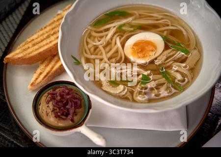Eine Schüssel warmer und wohltuender Nudelsuppe mit zarten Nudeln, einem weich gekochten Ei und herzhafter Brühe, begleitet von geröstetem Brot und einem schmackhaften Geschmack Stockfoto
