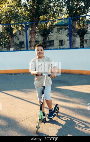 Junge reitet schwarz und grün Kick Scooter auf Outdoor Urban Court am Sommerabend, beiges T-Shirt und graue Shorts verspielter Kindheitsmoment gesund Stockfoto