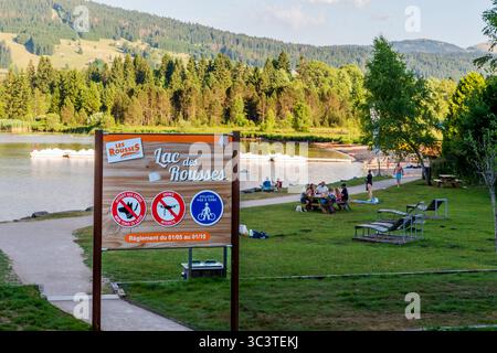 Les Rousses, Jura, Frankreich - 4. Juli 2025: LAC des Rousses Schild mit Menschen, die am See im Jura, Frankreich, entspannen. Stockfoto