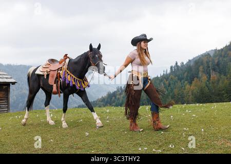 Eine junge Frau mit Cowboyhut und Jungs geht mit einem schwarz-weißen Pferd mit einem Sattel über ein grasbewachsenes Feld an einem bewölkten Tag in der Nähe eines Holzgebäudes. Stockfoto