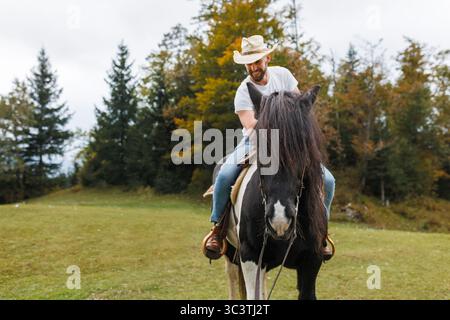 Ein Mann mit Cowboyhut reitet auf einem schwarz-weißen Pferd, während eine Frau in einem karierten Hemd lächelt und auf einem grasbewachsenen Feld mit ihm spricht. Stockfoto
