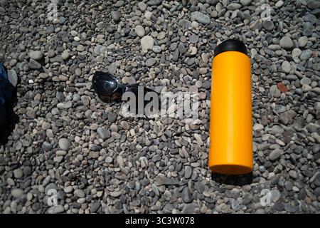 Eine erfrischende Sommerszene mit stilvoller Sonnenbrille und einer eleganten Wasserflasche auf glatten, sonnendurchfluteten Kieselsteinen am Rand eines ruhigen Strandes. Stockfoto