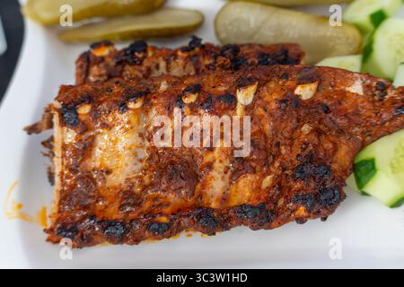 Winkelansicht Der Gegrillten Rippen. Barbecue-Rippchen mit einer verkohlten Kruste und Knoblauchstücken, die auf einem weißen Teller neben Gurken und frischen Gurkenscheiben ruhen. Zehn Stockfoto