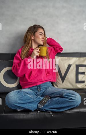 Ruhige junge Frau, die einen ruhigen Moment auf ihrem bequemen Sofa genießt, ein warmes Getränk genießt und eine Fernbedienung hält Stockfoto