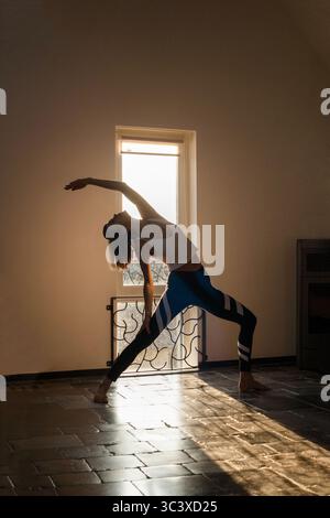 Schlanke, sportliche Frau, die zu Hause vor dem Fenster posiert, wo goldenes Sonnenlicht kommt. Yoga-Routine am Morgen, Selbstpflege, aktiver Lebensstil Stockfoto
