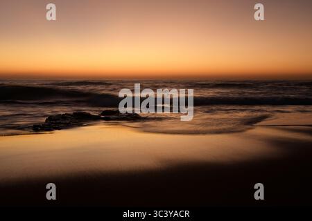 Wunderschöne Meereslandschaft mit brechendem Sonnenaufgang, stürzenden Wellen und goldenen Reflexen auf nassem Meersand, Winklespruit Beach, Amanzimtoti, Südafrika, Bewegungsunschärfe Stockfoto