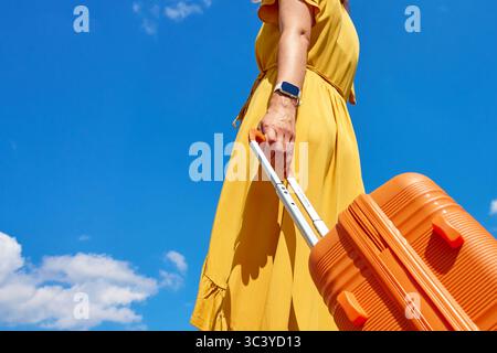 Frau in gelbem Kleid, die einen orangefarbenen Koffer vor blauem Himmel mit Wolken hält. Nahaufnahme einer Reisenden mit Gepäck. Konzept für stilvolles Reisen, va Stockfoto