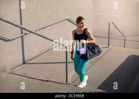 Die junge Frau in Activewear steigt die Treppe hinab, während sie eine Turntasche trägt. Sie verkörpert einen lebendigen Fitness-Lebensstil und setzt sich für Gesundheit und Wellness ein Stockfoto