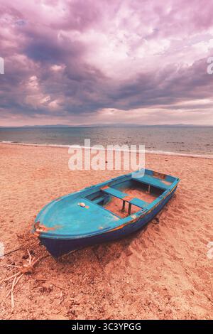 Verlassenes blaues Fischerboot am Sandstrand unter dramatischem bewölktem Himmel. Verwittertes Holzschiff in der Nähe des ruhigen Meeres, stimmungsvolle Atmosphäre mit rosafarbenen Sonnenuntergangstönen A Stockfoto