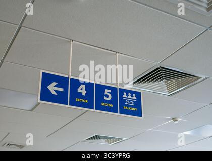 Ein blaues Schild mit weißen Pfeilen an der Decke in einem öffentlichen Bereich, das den Balkon, die Stände und die Sitzbereiche anzeigt. Stockfoto