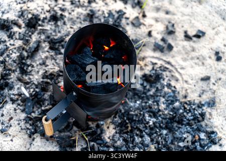 Nahaufnahme eines Holzkohlekamins mit glühenden Kohlen am Sandstrand - tragbares Feuerstartwerkzeug für umweltfreundliches Kochen im Freien Stockfoto