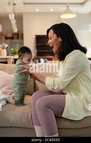 Mutter und Tochter halten das Baby in den Achselhöhlen und helfen ihr, auf dem Sofa mit rosafarbenen Kissen und Regalen zu stehen Stockfoto