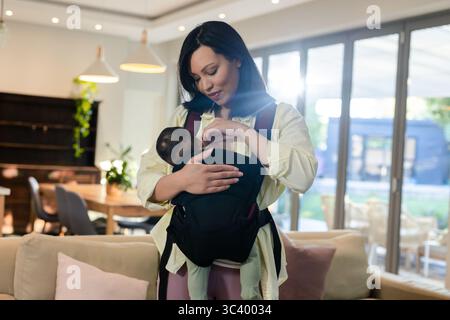 Asiatische Eltern und Kleinkinder, die die Babytragegurte vorne anpassen, in hellem Wohnzimmer mit Sofakissen Stockfoto