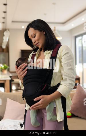Asiatische Mutter und Baby, die das Kind in einer nach vorne gerichteten Tragetasche mit beigefarbenem Sofa halten, Kopierraum Stockfoto