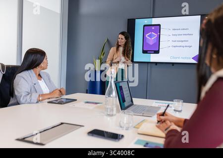 Verschiedene Geschäftsfrauen präsentieren Folien für die mobile Schnittstelle am Konferenztisch mit Laptops Tablets Stockfoto