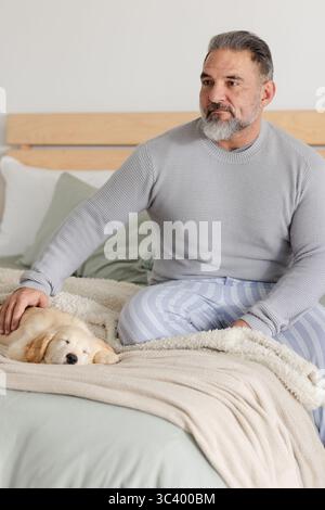 Mann mittleren Alters, der auf dem Bett sitzt und einen goldenen Welpen auf einer Decke unter dem Kopfteil streichelt, zu Hause, Kopierraum Stockfoto