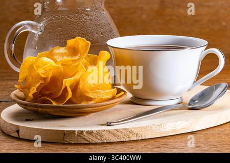 Nahaufnahme von süßen knusprigen Kartoffelchips auf Holzplatte mit einer weißen Keramikschale heißen Tees und einem Metalllöffel auf hölzernem Schneidebrett und einem Transpar Stockfoto