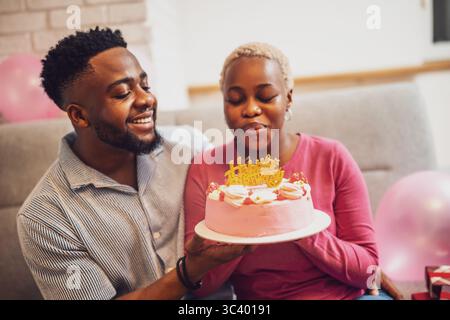 Glückliches junges Paar, das zu Hause den Geburtstag der Frau feiert. Sie halten Kuchen in der Hand und blasen die Geburtstagskerze. Stockfoto