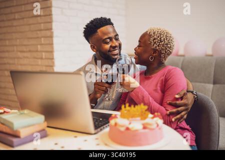 Glückliches junges Paar, das zu Hause den Geburtstag der Frau feiert. Sie toasten mit Wein und haben Videoanrufe mit der Familie. Stockfoto