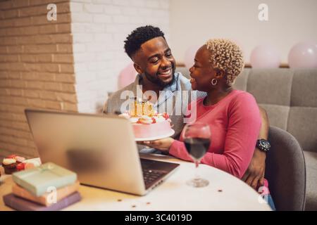 Glückliches junges Paar, das zu Hause den Geburtstag der Frau feiert. Sie halten Kuchen und haben Videoanrufe mit der Familie. Stockfoto